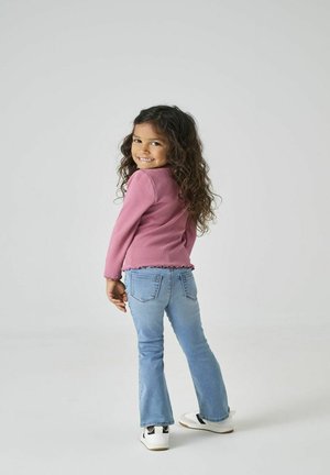 Lyserød bluse med lange ærmer og volangkant, kombineret med lyseblå, klokkeformede jeans. Børnene har hvide sneakers med sorte detaljer på.