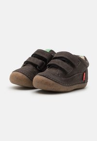 Zapatos de ante marrón con suelas de goma, que cuentan con correas de Velcro y un acento verde. Punta redondeada y detalles de costura en toda la superficie.