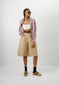 Beige weite Shorts mit einem Leopardenmuster-Gummibund, kombiniert mit einem weißen Cropped-Top und einem lilafarbenen Button-Up-Hemd. Die Accessoires umfassen weiße Sonnenbrillen und goldene Sneakers.