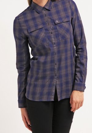 Femme portant une chemise à manches longues à carreaux bleus et gris avec des poches poitrine, associée à un pantalon noir, debout avec une main le long du corps.