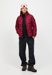 Bordeaux pufferjack met een gequiltde structuur, zwart ritstrui, marineblauwe cargo pants en bruine wandelschoenen. Lichtroze beanie.