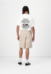 Tricou alb oversize cu text grafic negru pe spate, pantaloni scurți bej deschis și pantofi negri cu tălpi albe și șosete.
