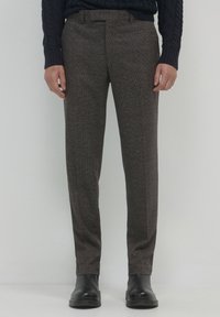 Mann in braunen karrierten maßgeschneiderten Hosen, schwarzen Lederstiefeln und einem dunklen marineblauen Strickpullover, der vor einem hellgrauen Hintergrund steht.
