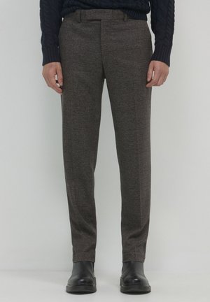 Homme portant un pantalon ajusté à carreaux marron, des bottes en cuir noires et un pull en mailles bleu marine, se tenant contre un fond gris clair.