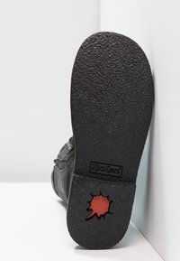 Semelle en caoutchouc noir avec une surface texturée, présentant une touche de rouge et le logo Kickers. Le côté de la chaussure est visible, montrant les détails de couture et de matériau.