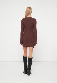 Pikkade blondide juustega naine, kellel on seljas pruun vertikaalribadega kootud kleit ja mustad kõrged kontsakingad, seisab valge tausta ees seljaga pöördunult.