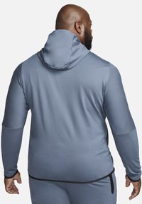 Soft, grey hoodie made of stretchy fabric. Features a fitted hood and long sleeves with textured accents. Includes a visible pocket and waistband.