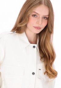 Witte oversized jas met een gladde, glanzende textuur, voorzien van grote borstzakken, een puntige kraag en zwarte drukknoopsluitingen.