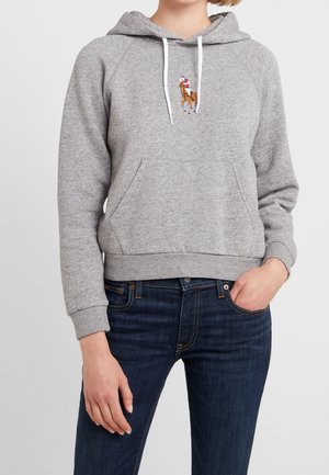 Persoon draagt een grijze hoodie met een kleine geborduurde afbeelding van een persoon op een paard, gecombineerd met donkerblauwe jeans en een hand in de zak.