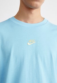 Ljusblå bomull T-shirt med en liten, broderad Nike-logotyp i gult på vänster bröstområde. Slät textur, rundad halsdesign.