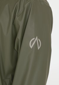 Olivgrüne, wasserdichte Jacke mit glatter Oberfläche. Sie verfügt über ein stilisiertes silbernes Logo am Ärmel und ein minimalistisches Design.