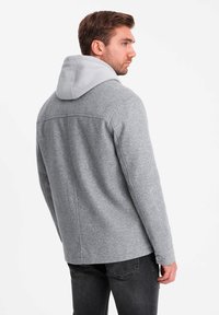 Veste en mélange de laine grise avec capuche gris clair, présentant une coupe droite, des coutures latérales et des poignets boutonnés. Finition texturée et design moderne.