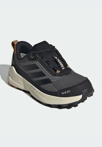 Chaussure de sport noire avec une tige en maille texturée, semelle en caoutchouc et accents gris. Présente un logo à trois rayures et un onglet de talon marron.