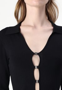Zwarte top met een diepe v-hals, voorzien van een knoopsluiting in het midden. Gemaakt van zachte stof met een kraag en lange mouwen.