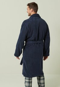 Tumši zils frotē auduma peldmētelis ar gariem piedurknēm, jostu viduklī un šaulu apkakli. Tekstūrēts audums, līdz ceļgaliem, valkāts ar kvadrātveida mēteļbiksēm.