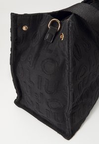 Borsa tote in tessuto nero con motivo a logo embossato, dotata di hardware dorato, una tracolla nera e una superficie testurizzata. Design con apertura superiore.