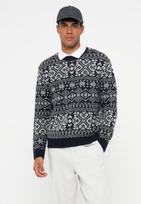 Sweter w marynarskim wzorze z białymi geometrycznymi wzorami, okrągły dekolt, długie rękawy i ściągacz na dole, zestawiony z białą bluzką z kołnierzykiem.
