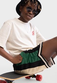 Crème T-shirt met korte mouwen en rode print, zwart-groene gestreepte shorts, zittend op een skateboard met rode wielen en een zwarte bucket hat.