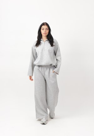 Kobieta idąca do przodu w szarej, oversize'owej bluzie i szerokich spodniach dresowych, w białych sneakersach na tle białej, gładkiej ściany.