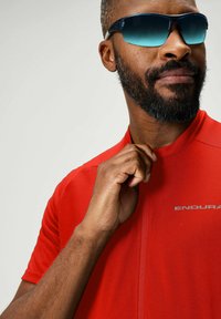 Rotes Sporttrikot mit hohem Kragen und kurzen Ärmeln, versehen mit einem reflektierenden silbernen Logo. Getragen mit dunklen Sonnenbrillen mit einem Verlaufstönung.