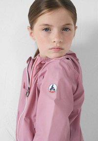 Lichtroze windjack met een staande kraag, ritssluiting aan de voorkant en elastische manchetten. Bevat een JOTT-logo op de linkermouw. Soepele textuur.
