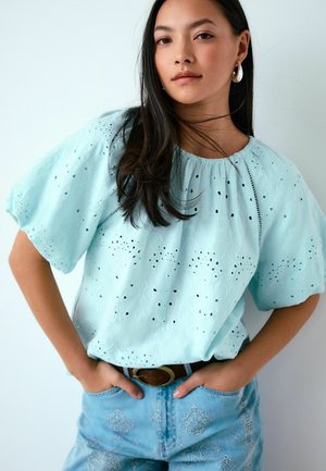 Noor naine pikkade tumedate juustega, kandes helesinist tikanditega pluusi, rõngaskõrvarõngaid ja mustrilisi teksapükse, käed taskutes, neutraalne taust.