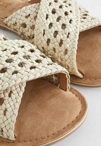 Geflochtene Sandalen aus cremefarbenem Leder mit offenem Zehenbereich, ausgestattet mit einem weichen Fußbett aus tanfarbenem Wildleder und geschwungenen Nähten an den Kanten.