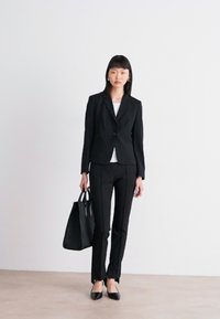 Crni krojeni blazer s matching hlačama, prilagođena silueta, uparen s bijelim topom, držeći crnu torbu i noseći crne balerinke.