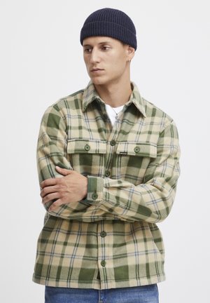 Flannel shirt in beige and green plaid, featuring two chest pockets, button-down front, and a classic collar. Knit navy beanie worn.