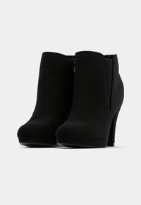 Botas de tobillo de ante negro con punta afilada, tacón alto y cierre de cremallera en el lado, con una textura lisa y mate.