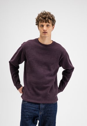 Jonge man met krullend haar, die een donkerpaarse langärmelig gestructureerd shirt en blauwe spijkerbroek draagt, staat met zijn handen op zijn heupen tegen een witte achtergrond.