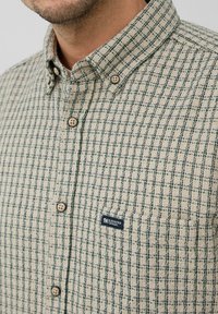 Man in een beige overhemd met knopen, met een groen ruitpatroon en een klein borstzakje met het label "Elevated Casual."
