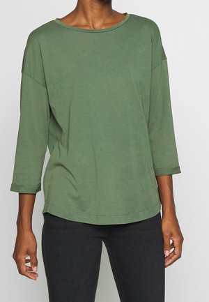 Persoon die een loszittend, olijfgroen shirt met driekwart mouwen en een zwarte broek draagt, zichtbaar van nek tot heupen tegen een effen achtergrond.