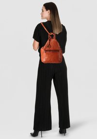 Sac à dos en cuir marron avec une texture lisse, un sommet arrondi et un logo circulaire décoratif. Dispose d'une poche zippée à l'avant et d'une sangle réglable.