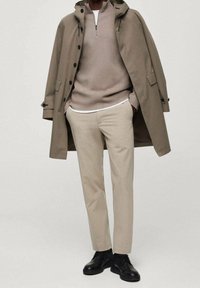 Hombre vestido con pantalones beige, suéter de medio cierre en color topo, trench coat topo sobre los hombros, y zapatos negros de cordones.