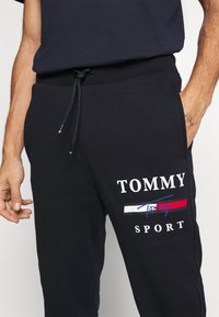 Schwarze Jogginghose mit Kordelzugbund; verfügt über ein weißes "TOMMY SPORT"-Logo und einen farbigen Akzentstreifen in Rot und Blau am Oberschenkel.