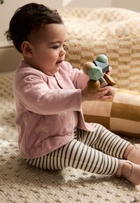 Spædbarn iført en lyserød tekstureret cardigan, der holder flerfarvede træklodser. Stribede leggings, lyserøde sko. Indendørs tekstureret baggrund.