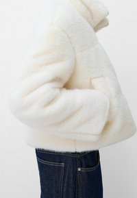 Veste en fausse fourrure blanche avec un design rectangulaire, un col haut et des poches latérales. Associée à un jean en denim foncé avec une poche arrière classique.