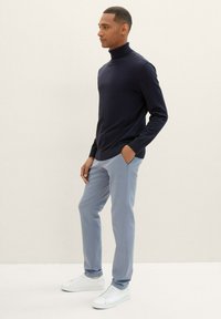 Navy turtleneck sweater with ribbed texture, paired with light blue slim-fit trousers and white sneakers. Standing profile against a neutral background.