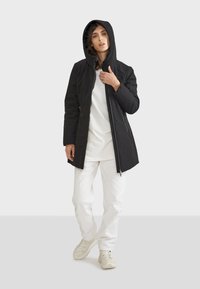 Zwarte gewatteerde jas met een capuchon, voorzien van een voorkleur en een slim fit. Draagt over een witte top en lichte broeken, met een soepele textuur.
