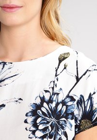 Top floreale con base bianca, caratterizzato da motivi floreali blu e oro, tessuto morbido, scollatura rotonda e maniche corte con dettagli neri.