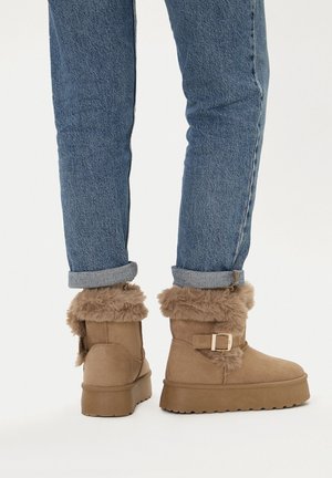 Beżowe futrzane kozaki z solidną podeszwą, wyposażone w pasek z klamrą i miękką fakturą, zestawione z podwiniętymi niebieskimi dżinsami.