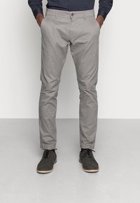 Šedé chino kalhoty se slim fit, předními kapsami a zapínáním na knoflík. Nosí se s tmavými botami s bílou podrážkou.
