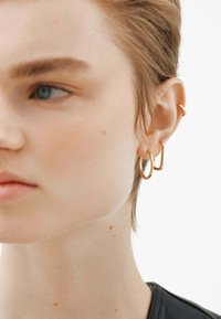 Pendientes de aro y de botón de oro llevados en la oreja, con formas variadas; textura suave contra el cabello corto y tono de piel clara, fondo minimalista.