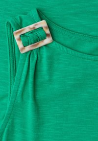 Vêtement en tissu vert avec une boucle rectangulaire en écaille de tortue rassemblée à l'encolure, montrant la texture et les détails des coutures.