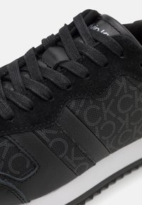 Black sneaker with a mix of textured leather and smooth synthetic material. Features prominent logo pattern, flat laces, and rubber sole.