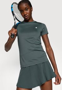 Pilkos spalvos sportinė marškinė su trumpomis rankovėmis ir apvalia iškirpte, derinami su atitinkama platėjančia sijona. Modelis laiko teniso raketę.