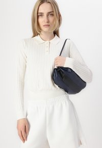Camisa polo de manga larga blanca con textura acanalada, tres botones y cuello crema, combinada con pantalones cortos blancos de cintura elástica y un bolso de cuero negro.
