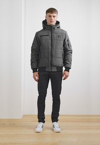 Polstret sort jakke med lynlås foran, hætte og elastiske manchetter; har to sidelommer og en brystlomme. Båret med mørke bukser og hvide sneakers.