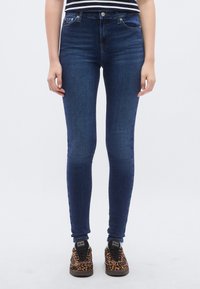 Sötétkék farmerból készült szűk szabású nadrág, közepes derekú, ötlovas zsebekkel, sima textúrával. Fekete és leopárdmintás sportcipővel párosítva.